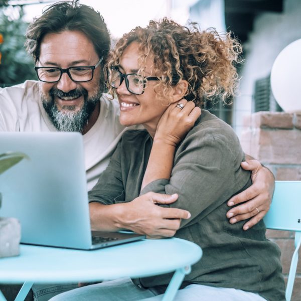 Modern couple enjoying video call on laptop sitting outside home in the garden. Wireless connection. People man and woman using computer outdoor enjoying time and love. Man embracing woman bonding